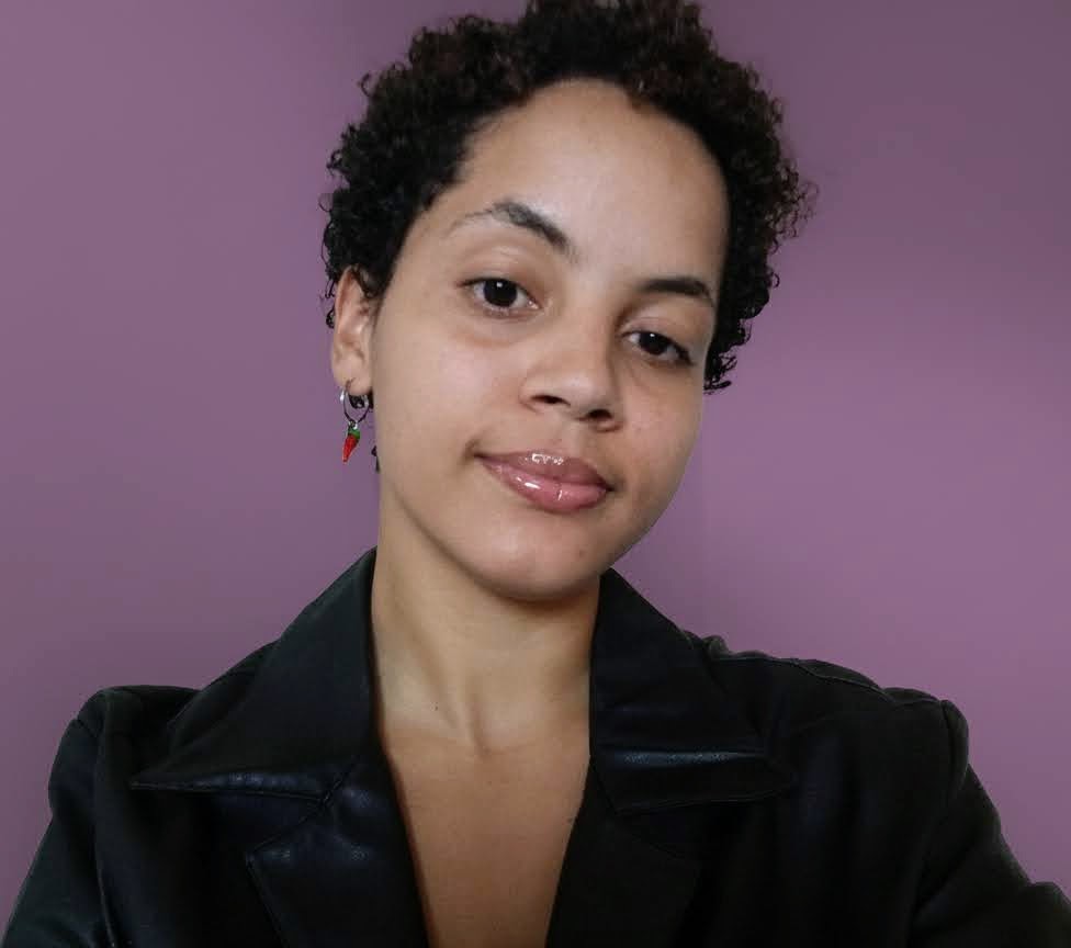 Mulher baixa, parda, cabelo curto e cacheado, olhos castanhos e veste uma jaqueta preta